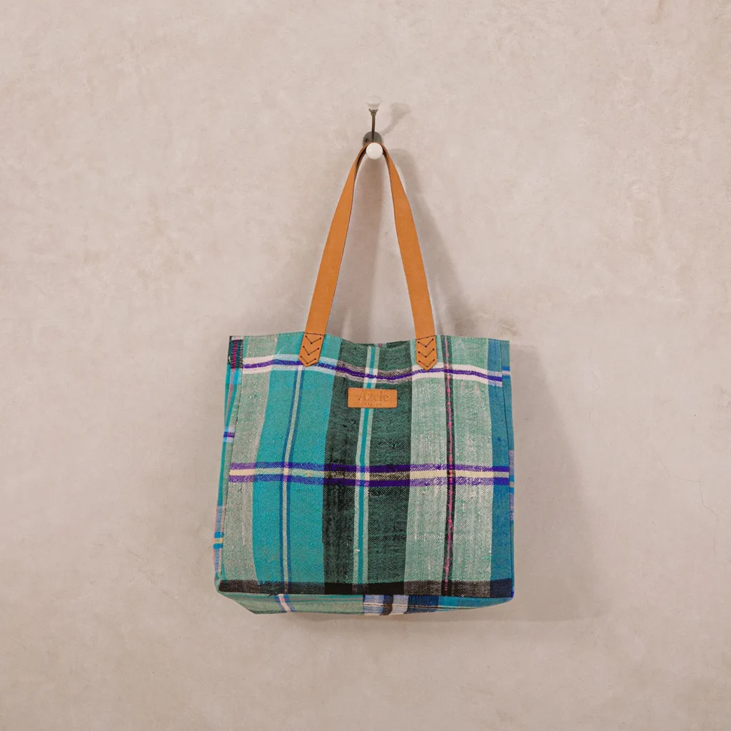 A plaid tote bag with green and blue tones, featuring brown leather handles, hangs on a simple wall hook against a light beige wall.