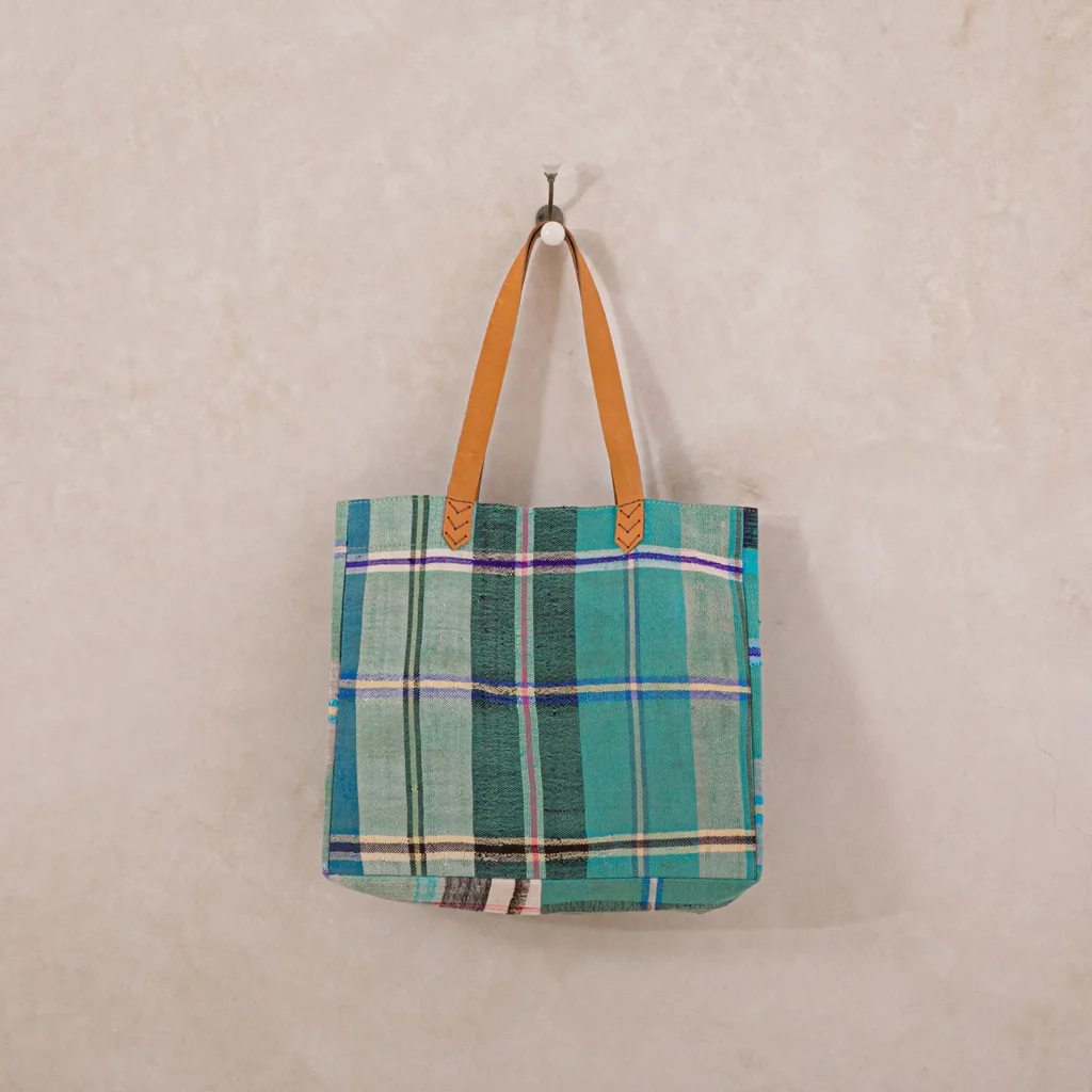 A plaid tote bag with brown leather straps hangs on a wall hook.