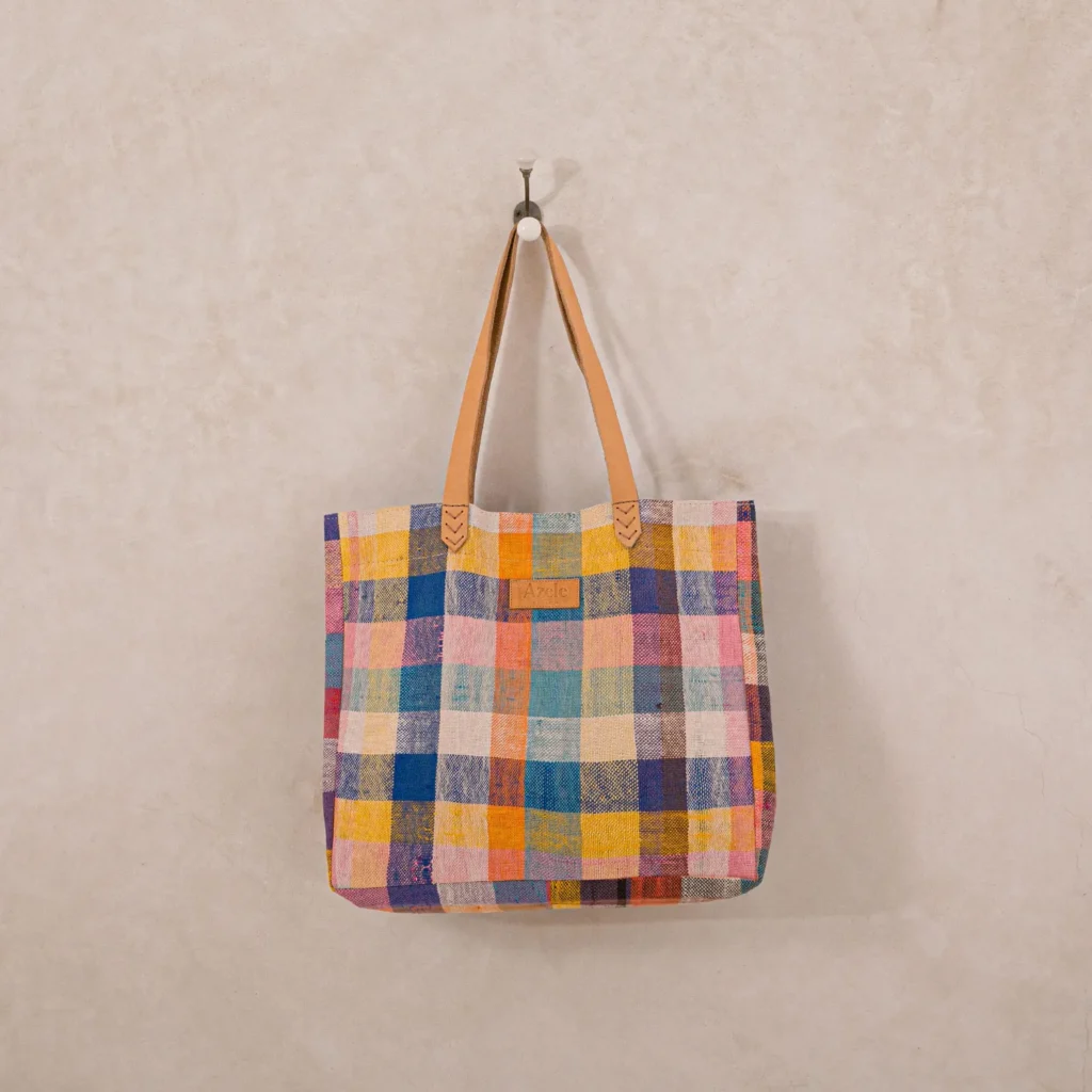 An upcycled shopper bag with a multicolored checkered pattern and tan leather handles hangs on a wall hook.