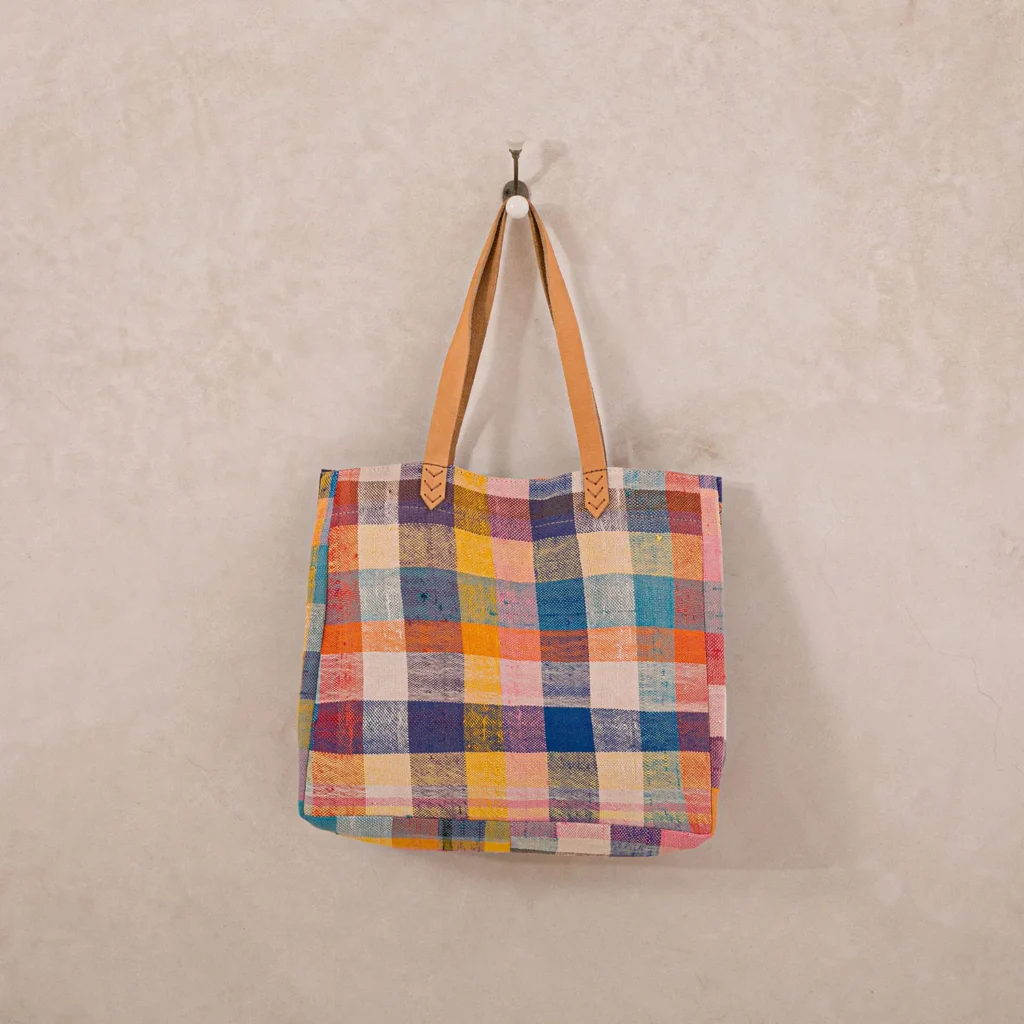 A multicolored plaid tote bag with tan leather handles hangs on a wall hook.