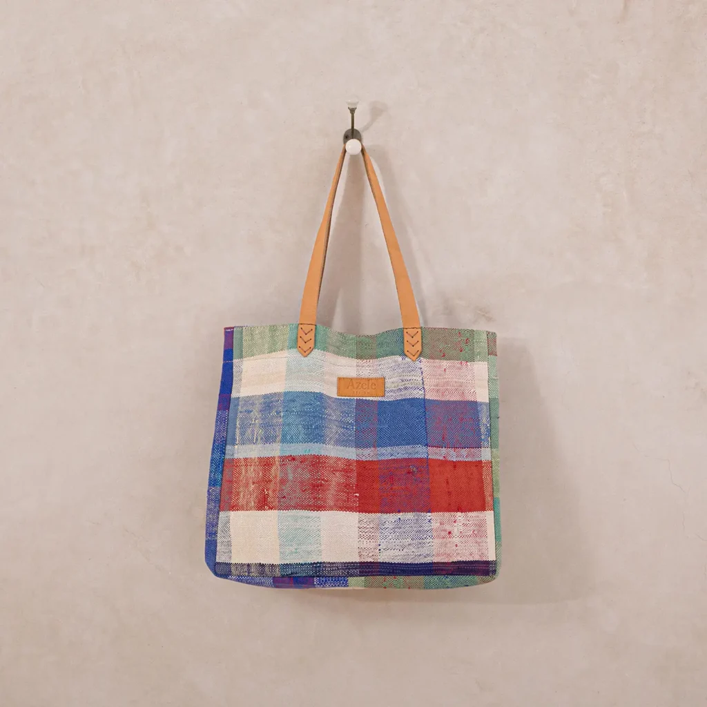 A multicolored checkered bag with tan leather handles hangs on a hook against a plain white wall.