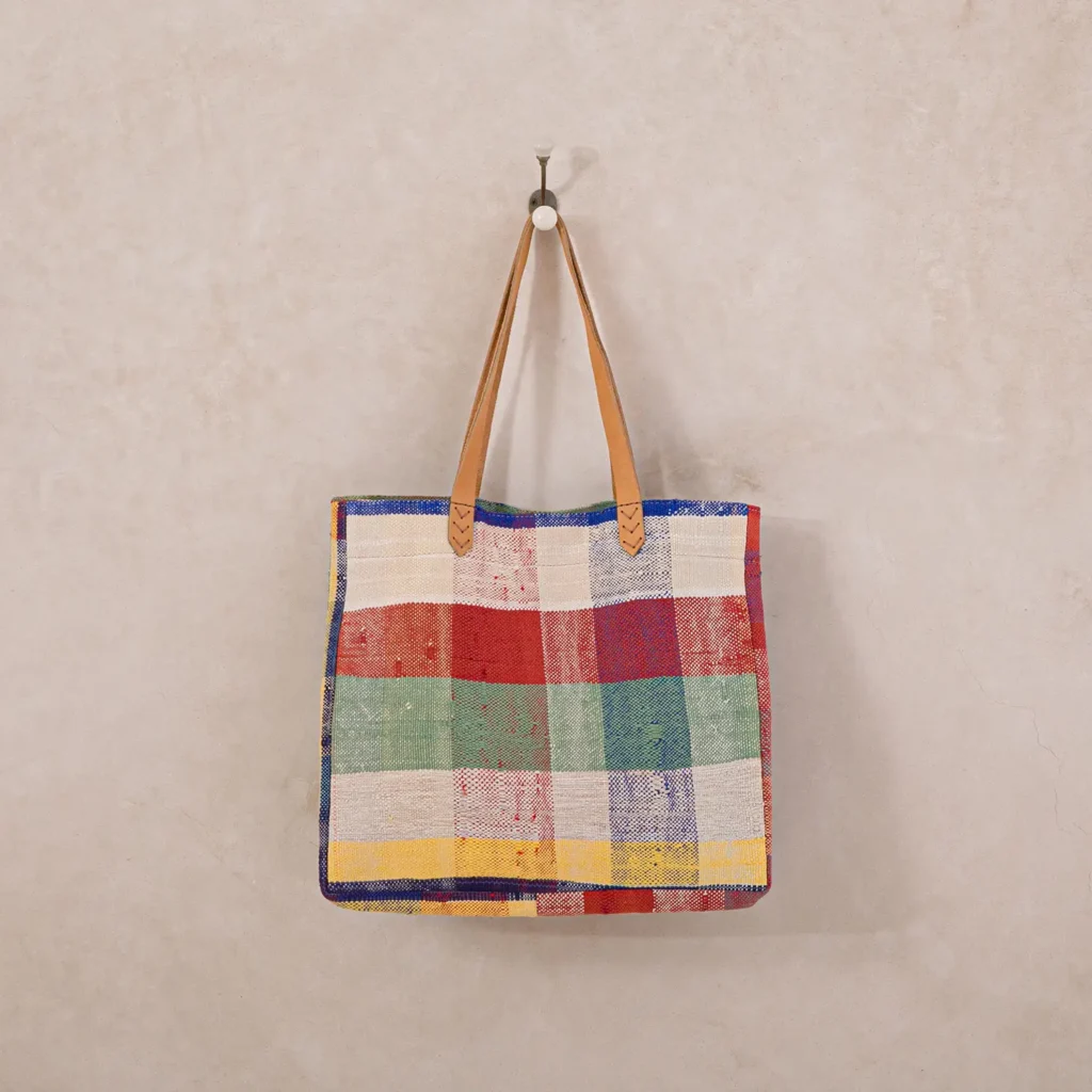 A multi-colored plaid tote bag with brown leather handles hangs on a wall hook against a textured cream background.