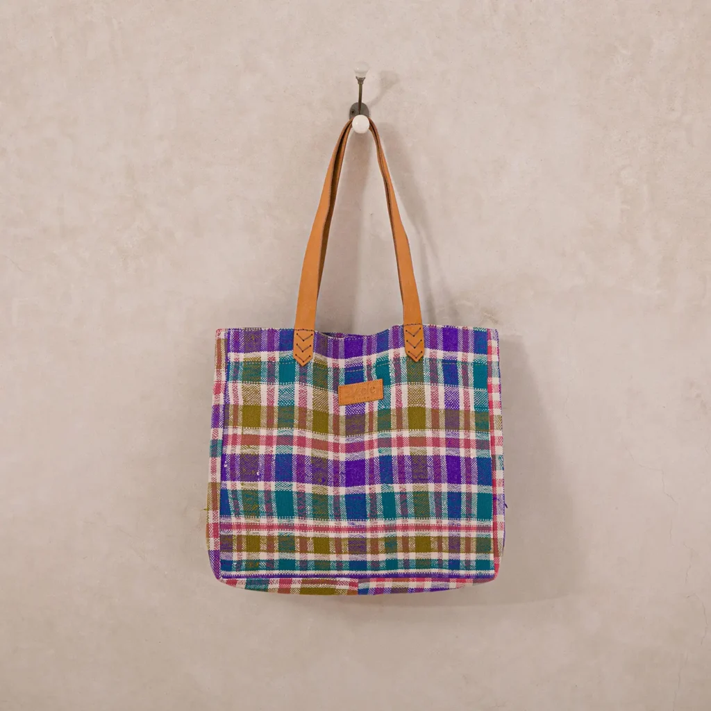 A plaid tote bag with brown handles hangs on a wall hook. The bag features a checkered pattern in purple, blue, green, and pink.