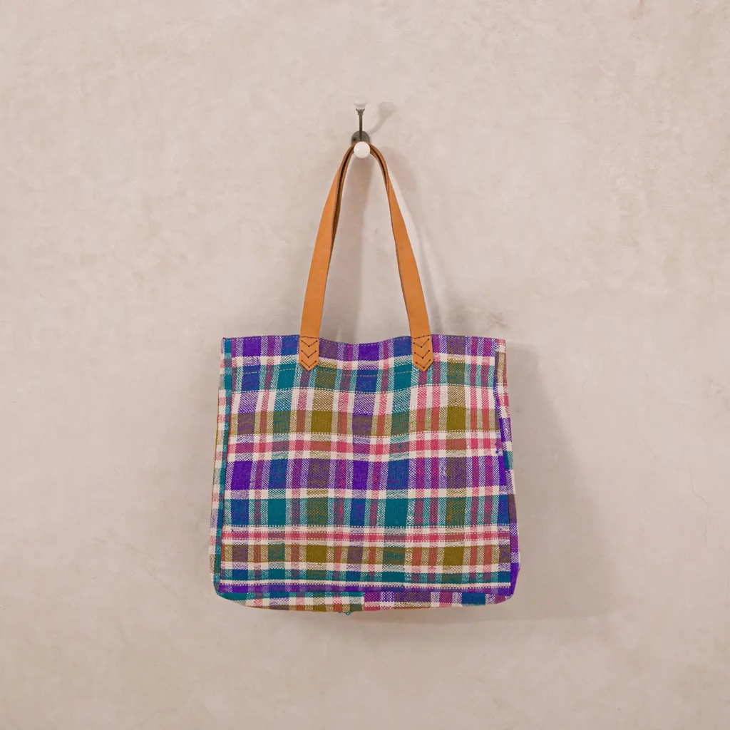 A plaid tote bag with a multicolored pattern in purple, green, and red hangs on a single hook against a beige wall. It has brown leather straps.