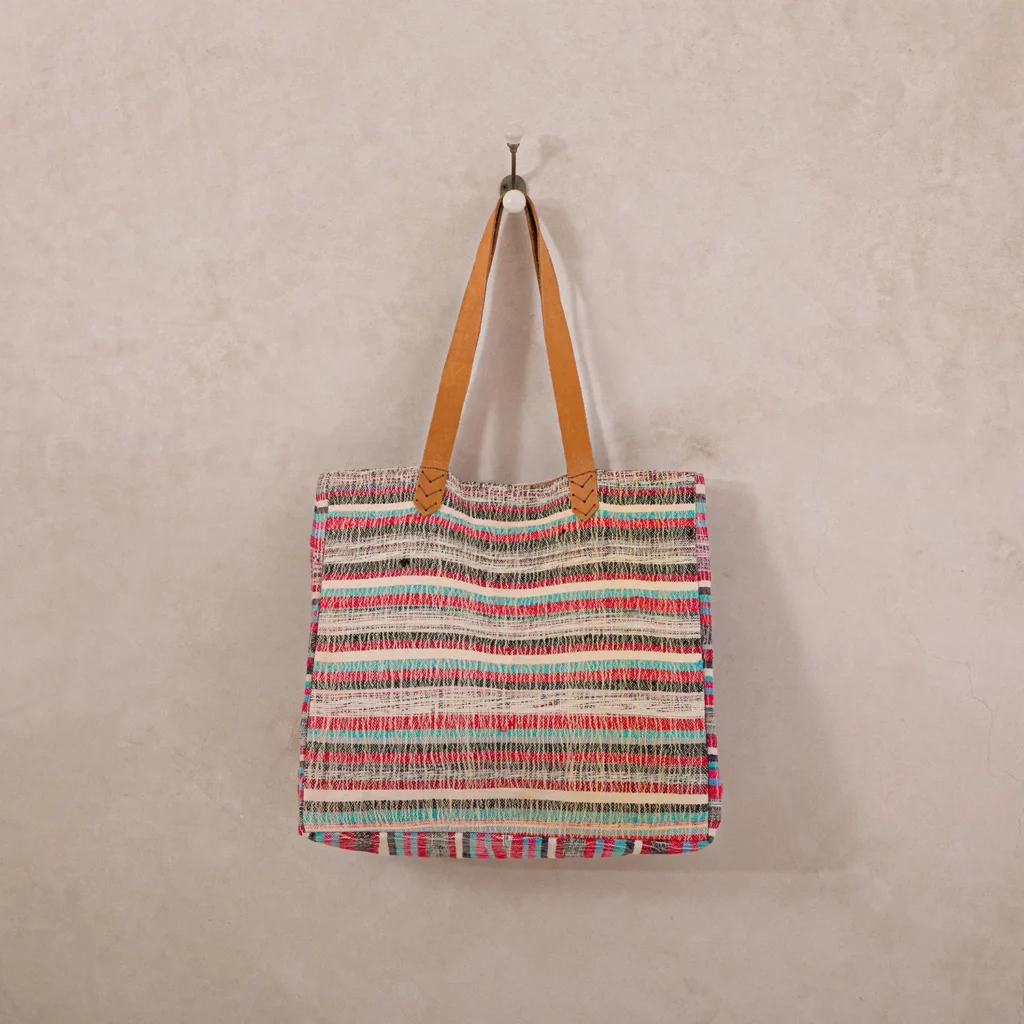 A multicolored striped woven tote bag with brown handles hangs on a metal hook against a plain beige wall.