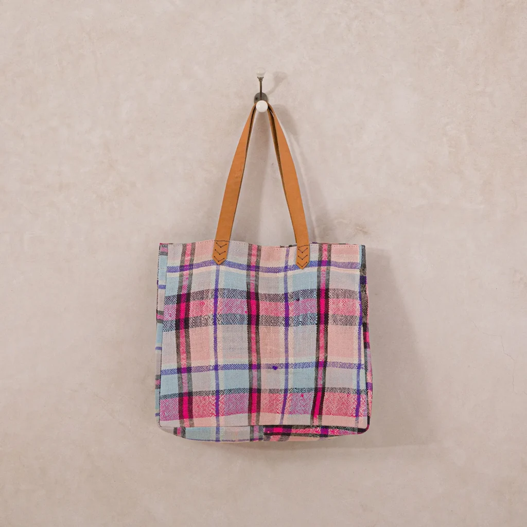 A plaid tote bag with pink, blue, and beige squares hangs on a wall hook. The bag has brown leather handles.