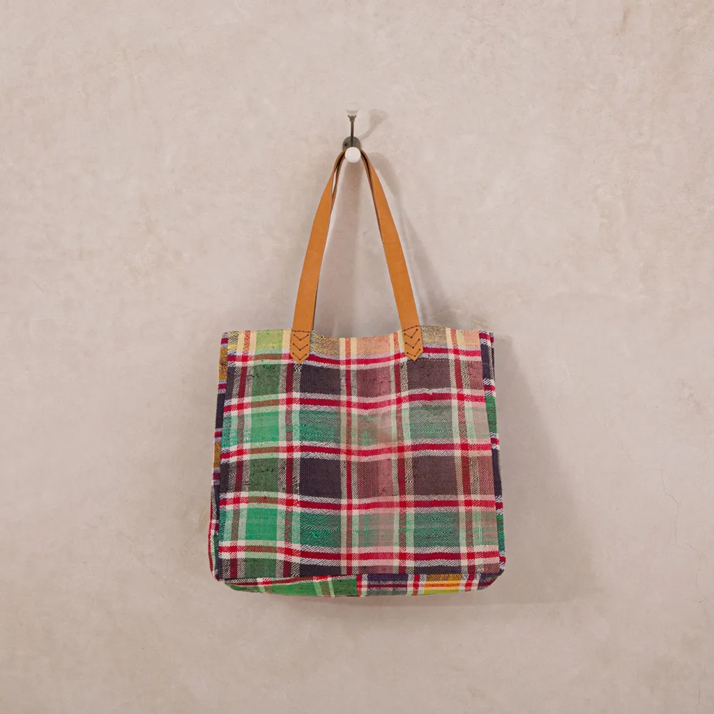 Plaid tote bag with red, green, and brown patterns and tan leather handles hanging on a white wall hook.