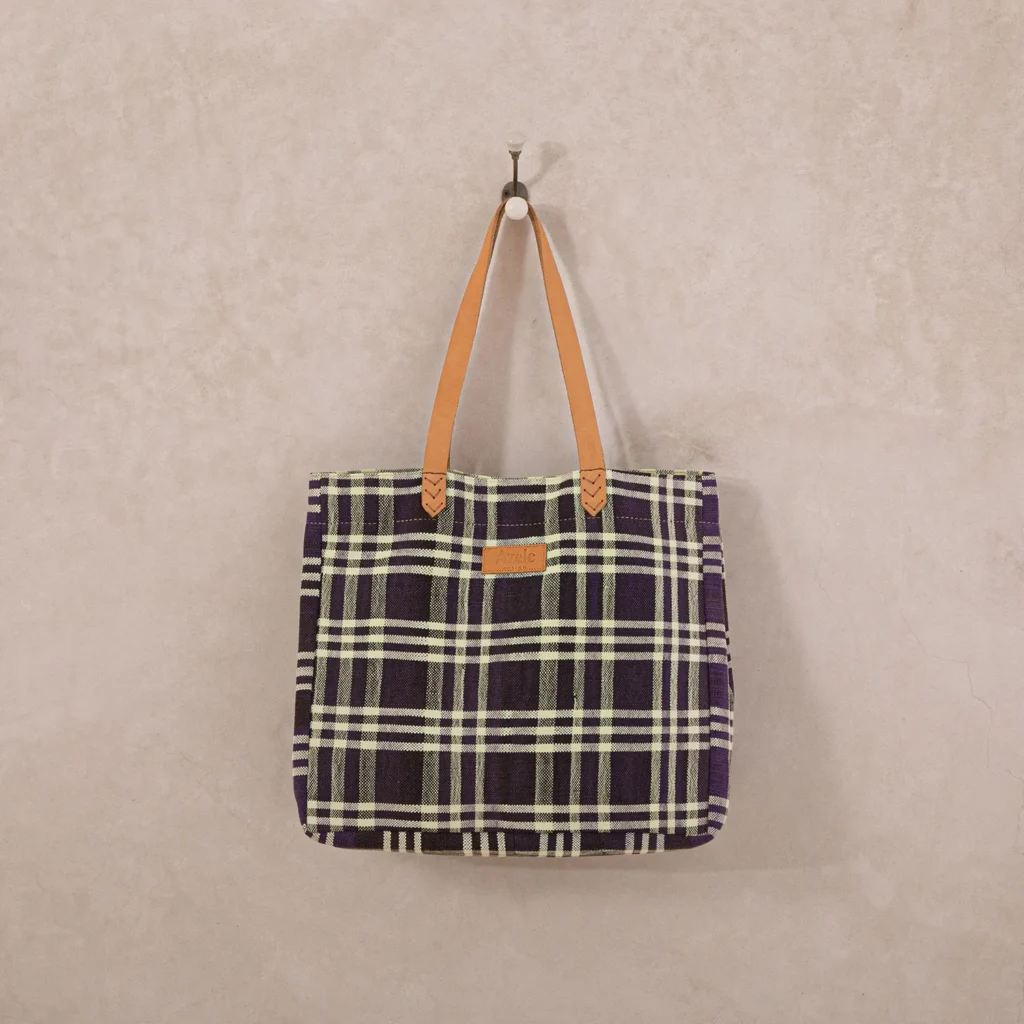 Plaid tote bag with beige and black checkered pattern, brown leather straps, hanging on a wall hook.