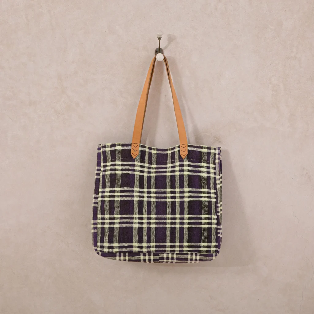 Plaid tote bag with black, white, and green pattern, hanging on a wall hook. It has brown leather handles.