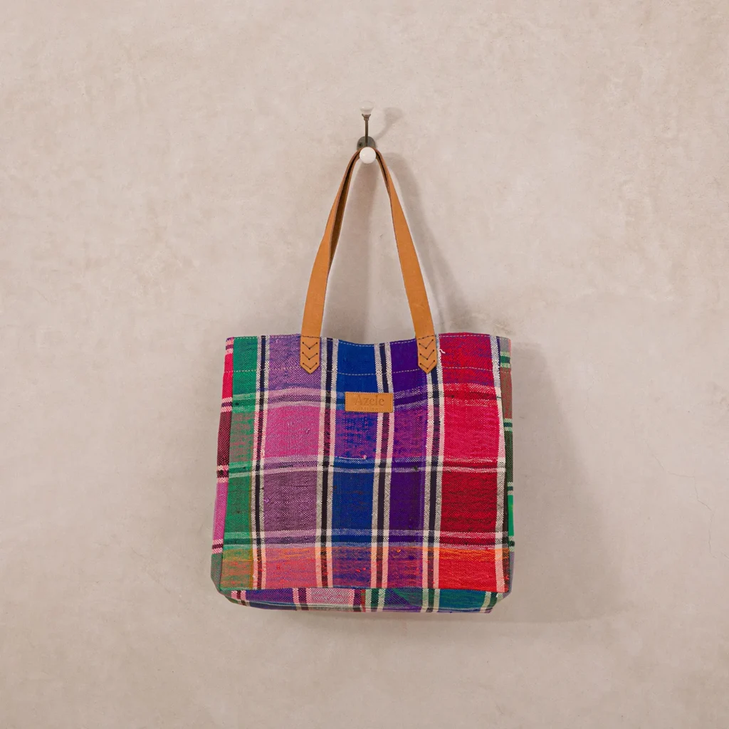A multicolored plaid tote bag with brown leather handles hangs on a hook against a plain beige wall.