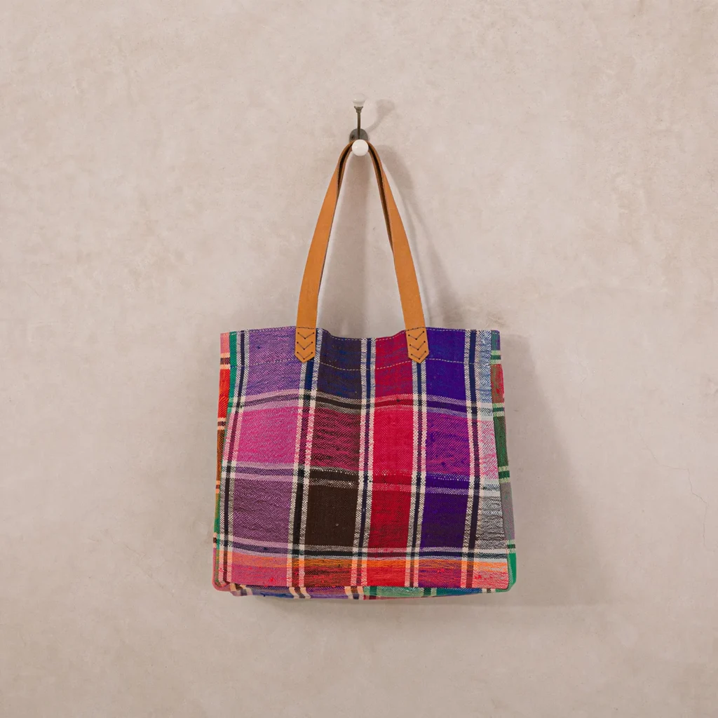 A colorful, plaid-patterned tote bag with brown leather handles hangs on a wall hook.