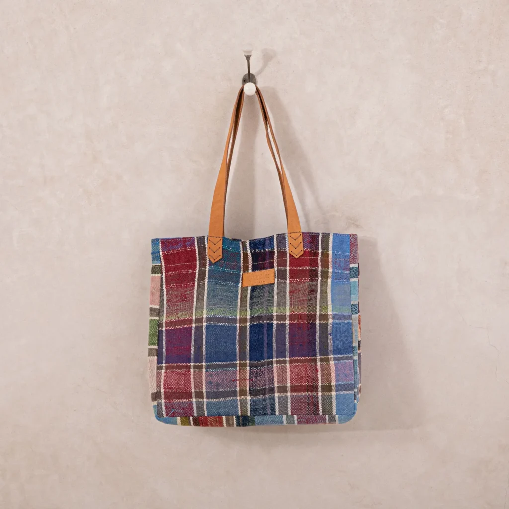 An upcycled shopper bag, featuring a plaid patchwork design with brown leather straps, hangs elegantly on a wall hook.
