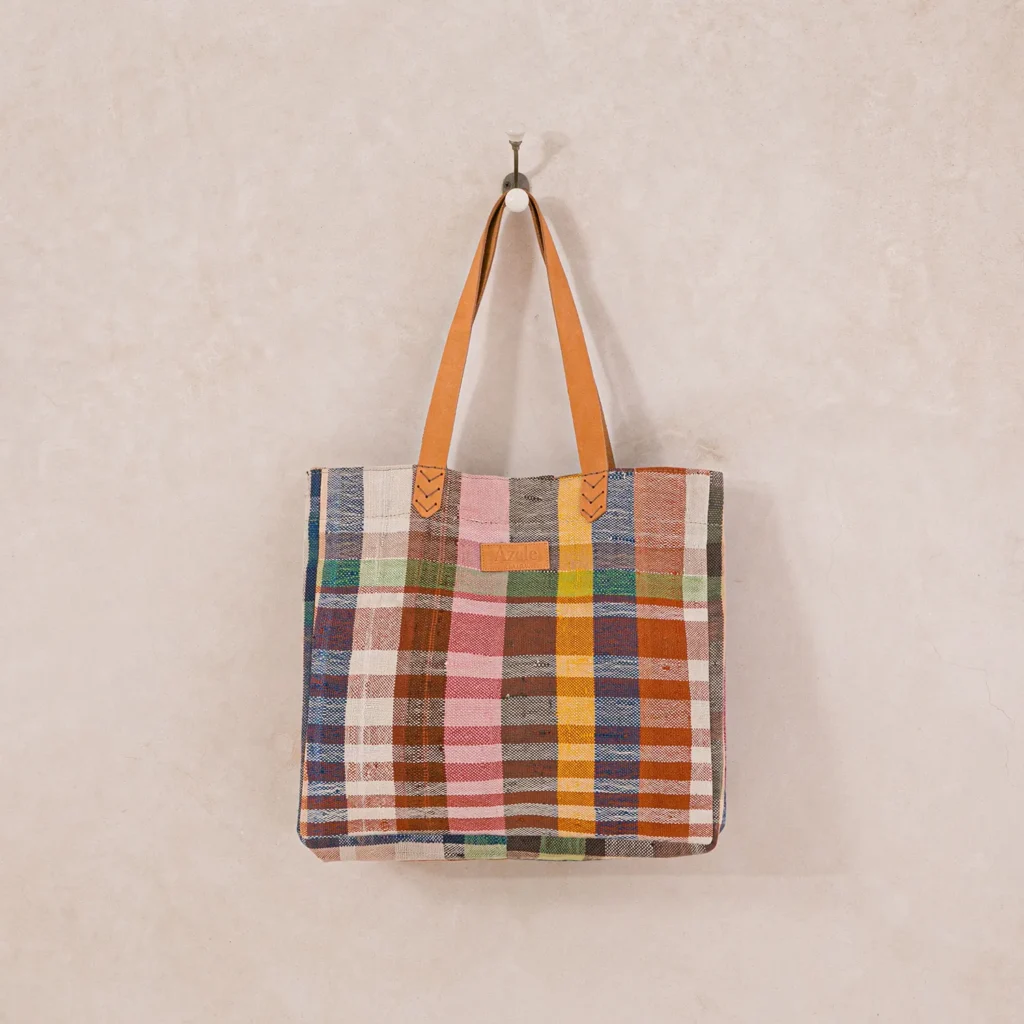 A multicolored plaid tote bag with tan leather handles hangs on a hook against a light beige wall.