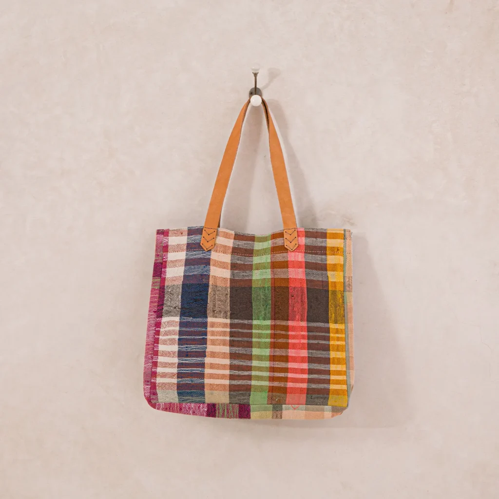 A multicolored plaid tote bag with brown leather handles hangs on a wall hook.