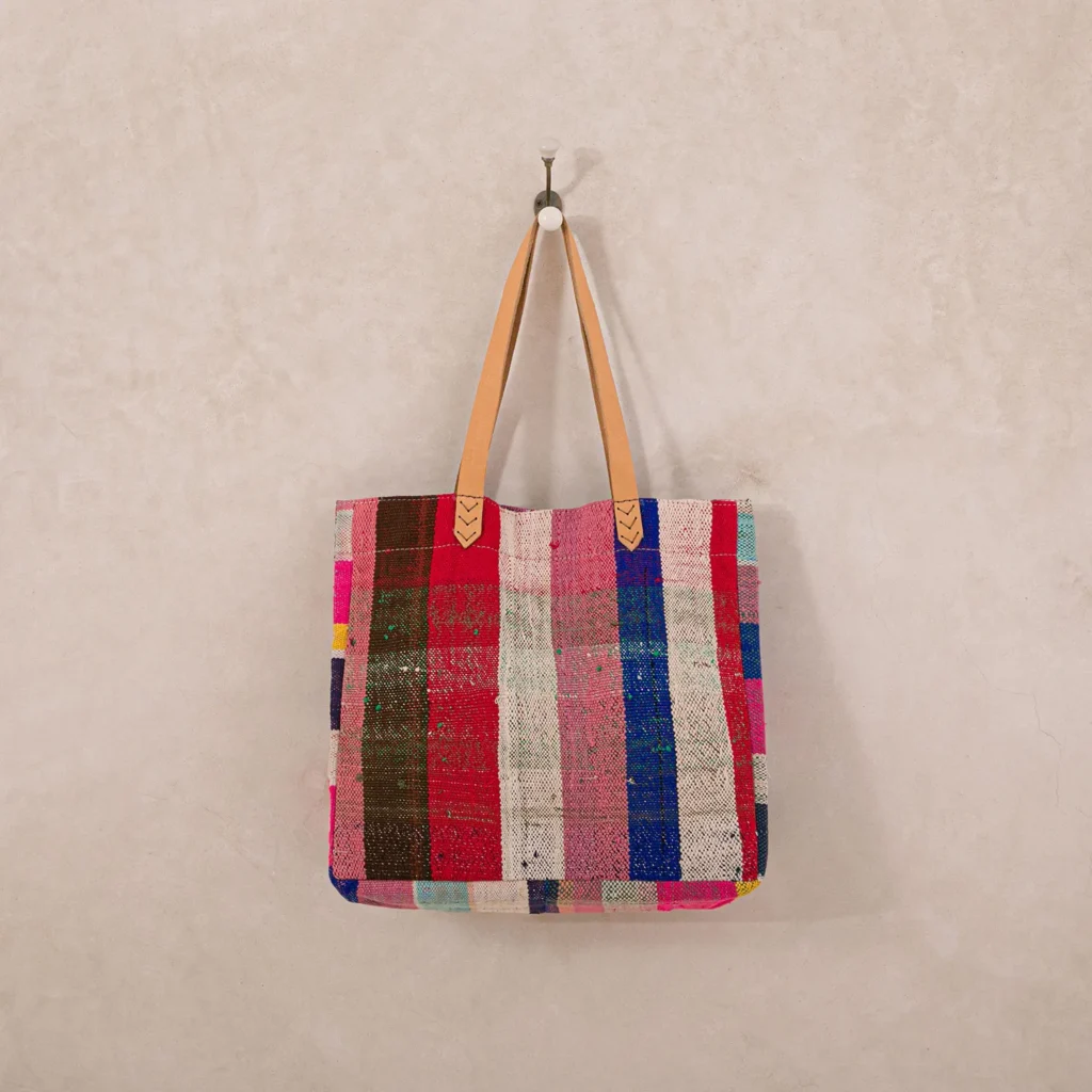 An upcycled shopper bag, featuring multicolored stripes and tan handles, hangs elegantly on a wall hook.