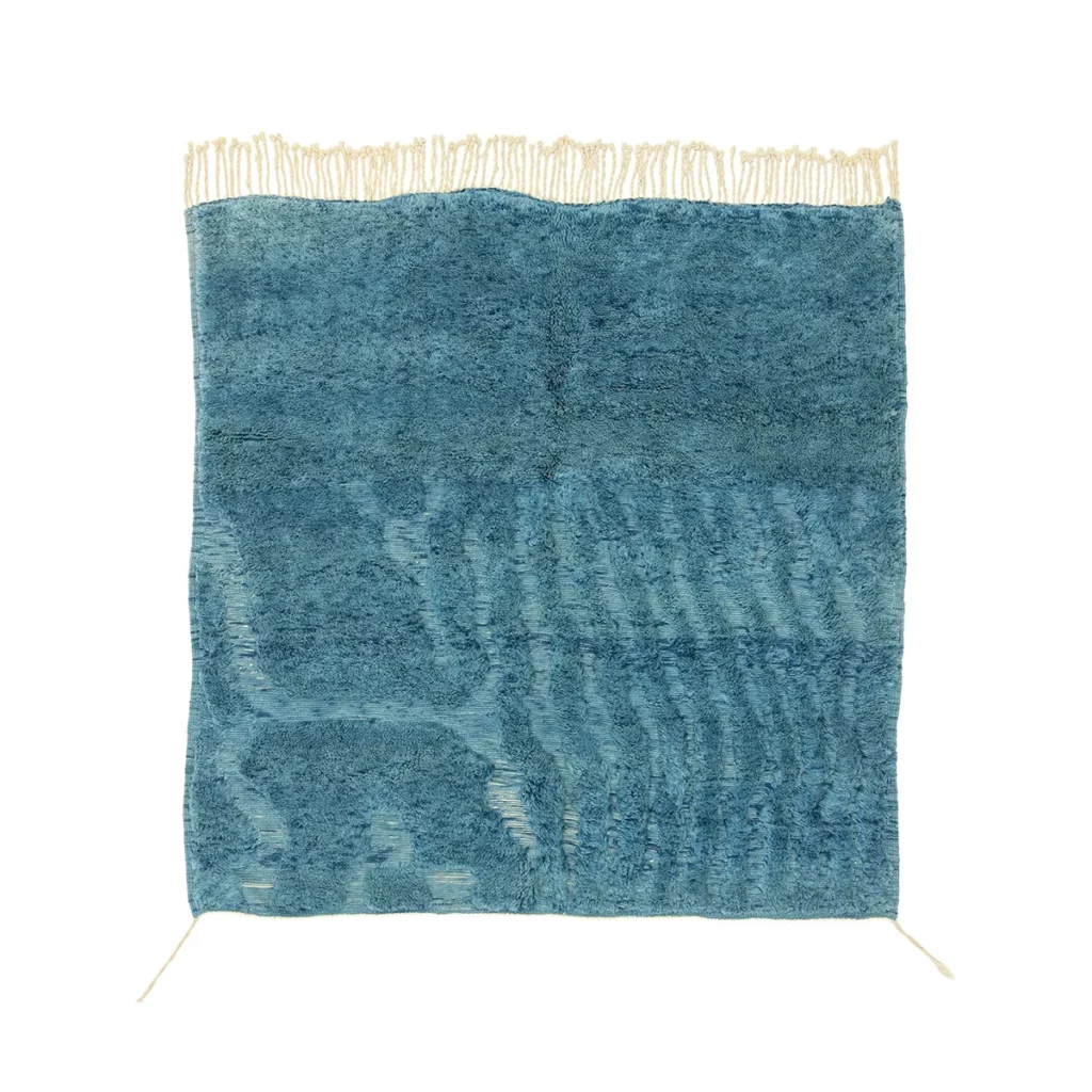 This handmade home decor piece features a blue textured rug with irregular patterns and fringe along the top edge, adding a touch of elegance to any space.