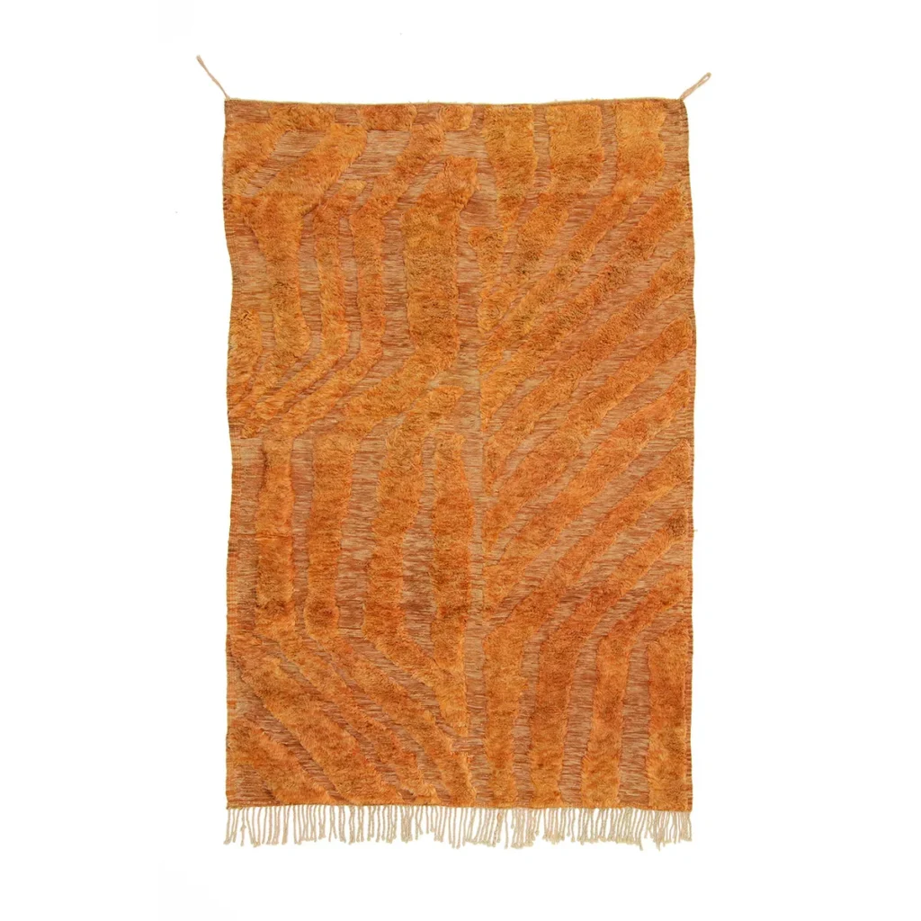 A luxurious, large orange rug with abstract zigzag patterns and fringe at the bottom edge hangs against a plain white background, showcasing exquisite craftsmanship in handmade home decor.