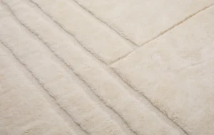A close-up view of an artisanal rug showcases its soft, cream-colored texture with subtle geometric patterns created by varying pile heights, reflecting the intricate craftsmanship typical of authentic Moroccan rugs.