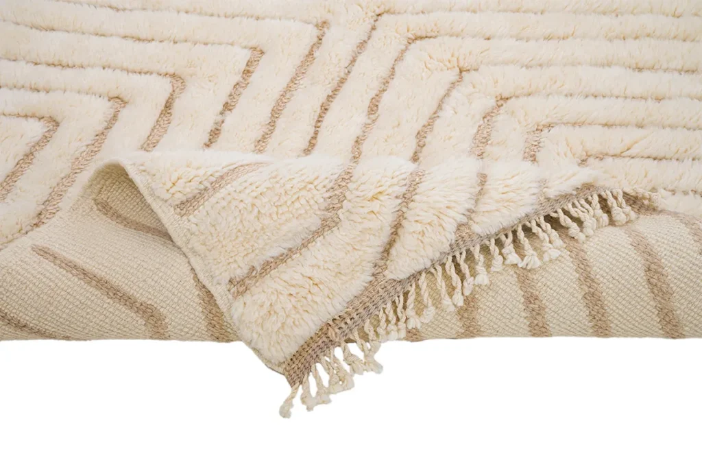 This artisanal rug, featuring a beige and cream pattern with fringed tassels on two sides, is partially folded to reveal the intricate underside.