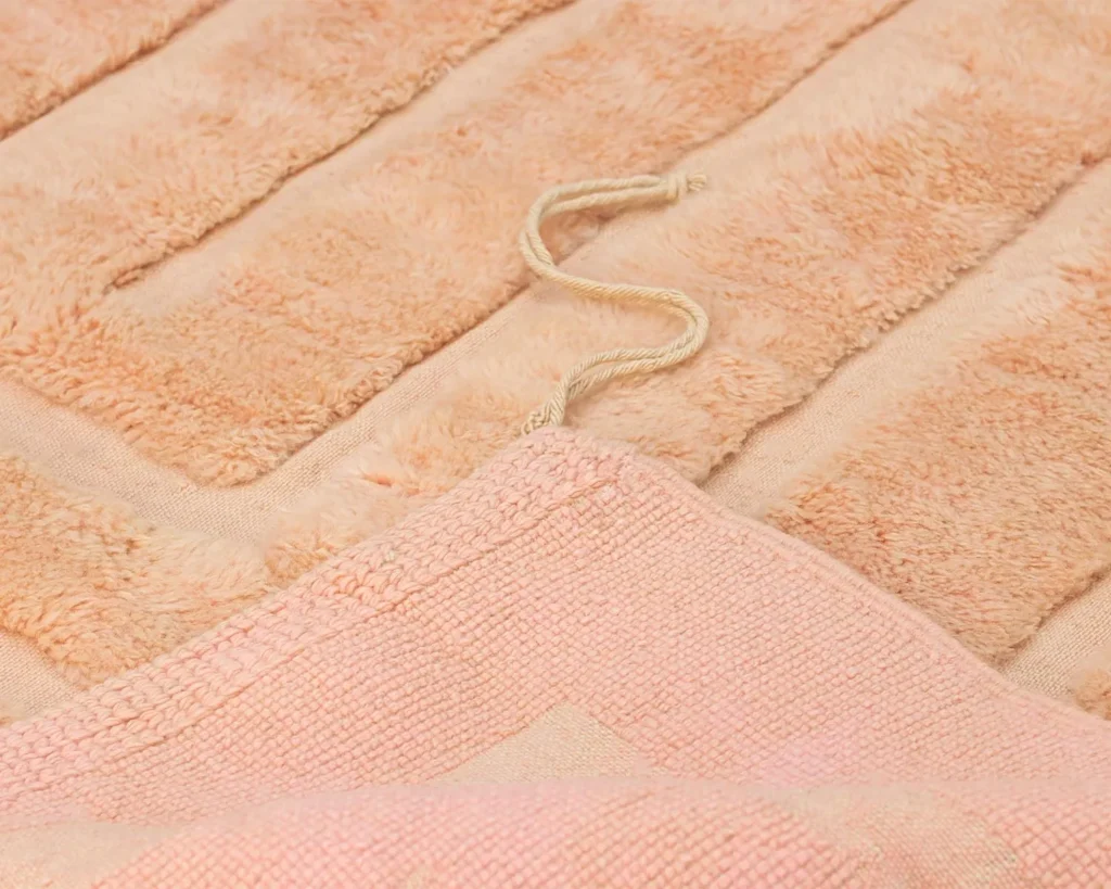 Close-up of a peach-colored, textured luxury rug with a looped cotton cord on top. The piece features a plush surface and ribbed pattern, blending elegance with comfort seamlessly.