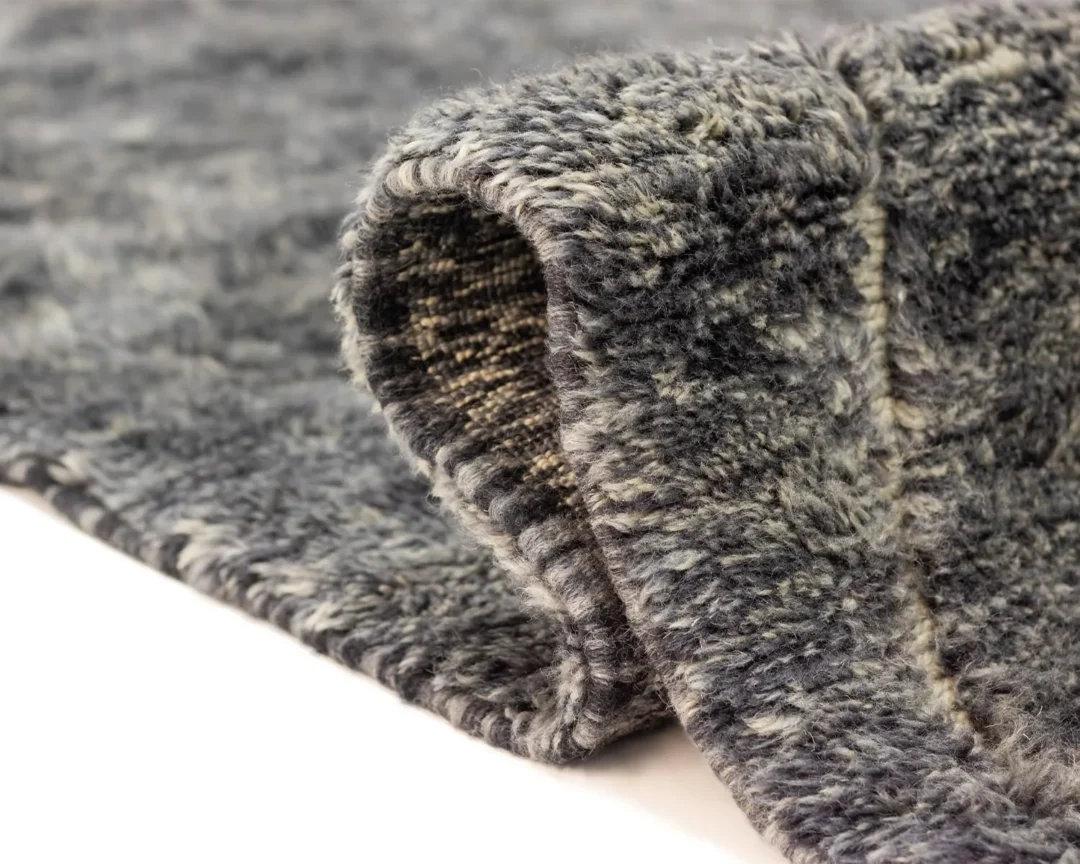Close-up of an artisanal rug with a curled edge, showcasing the soft texture and woven underside. This hand-knotted piece displays a mix of light and dark gray shades, creating a mottled appearance that adds depth to any space.