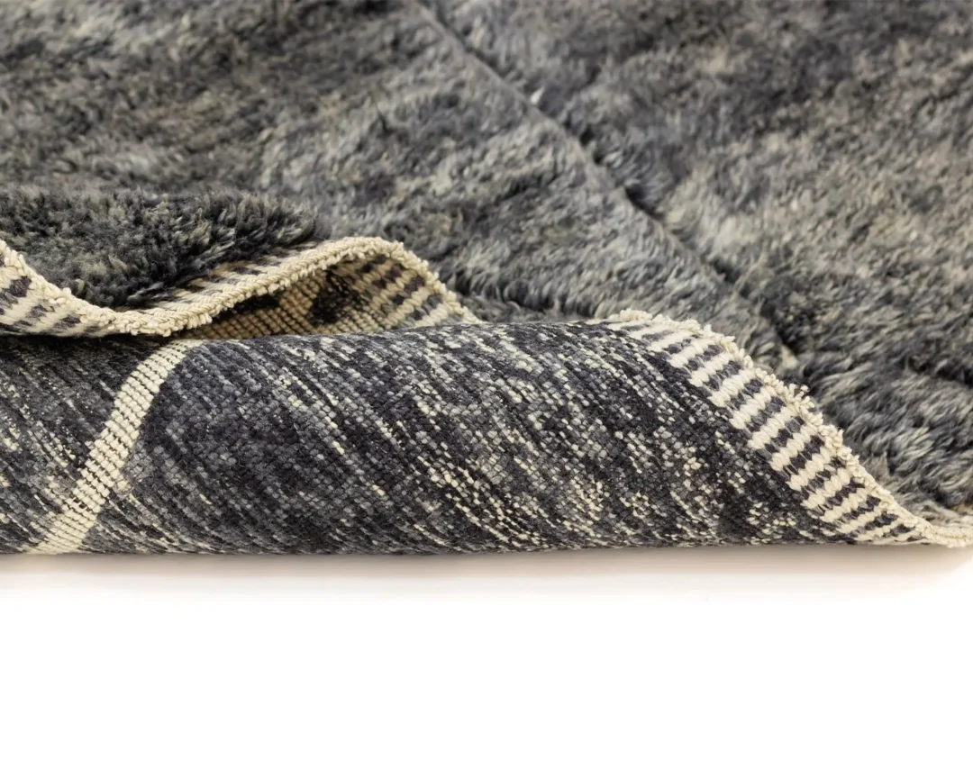 A close-up of an artisanal gray textured rug with one corner folded over, revealing a striped backing.