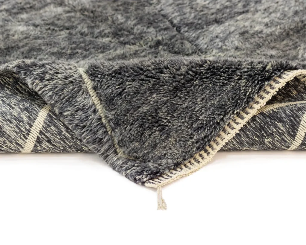 This close-up reveals the intricate beauty of an authentic Moroccan rug. It boasts a gray, hand-knotted plush upper layer and a patterned underside accented by tassel edging, showcasing artisanal craftsmanship.