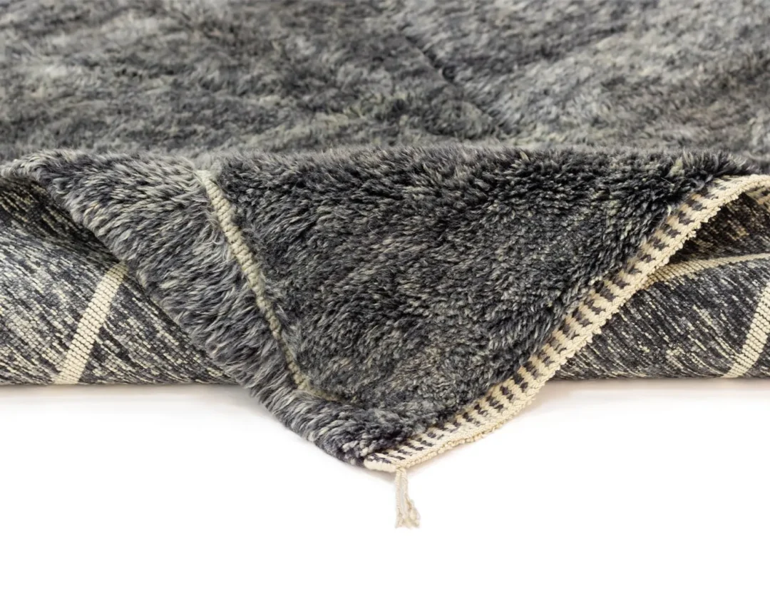This close-up reveals the intricate beauty of an authentic Moroccan rug. It boasts a gray, hand-knotted plush upper layer and a patterned underside accented by tassel edging, showcasing artisanal craftsmanship.