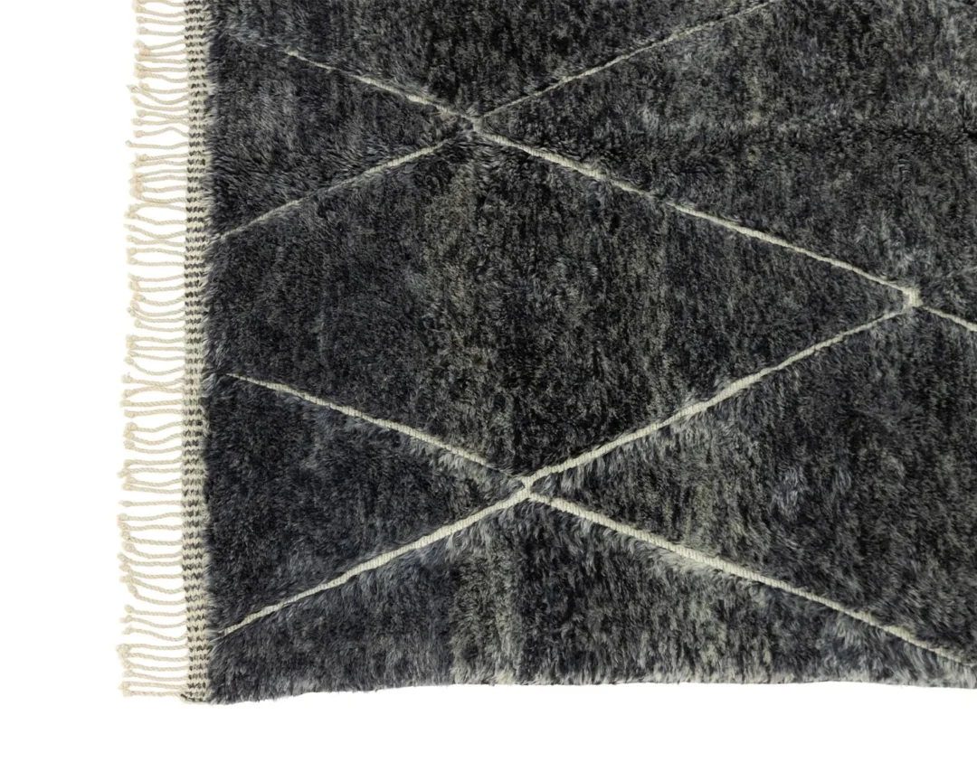 The dark gray plush rug features white geometric lines that form diamond patterns, and its fringed edge on the left adds a touch of elegance. This artisanal rug exudes the charm of authentic Moroccan craftsmanship, making it a unique addition to any space.