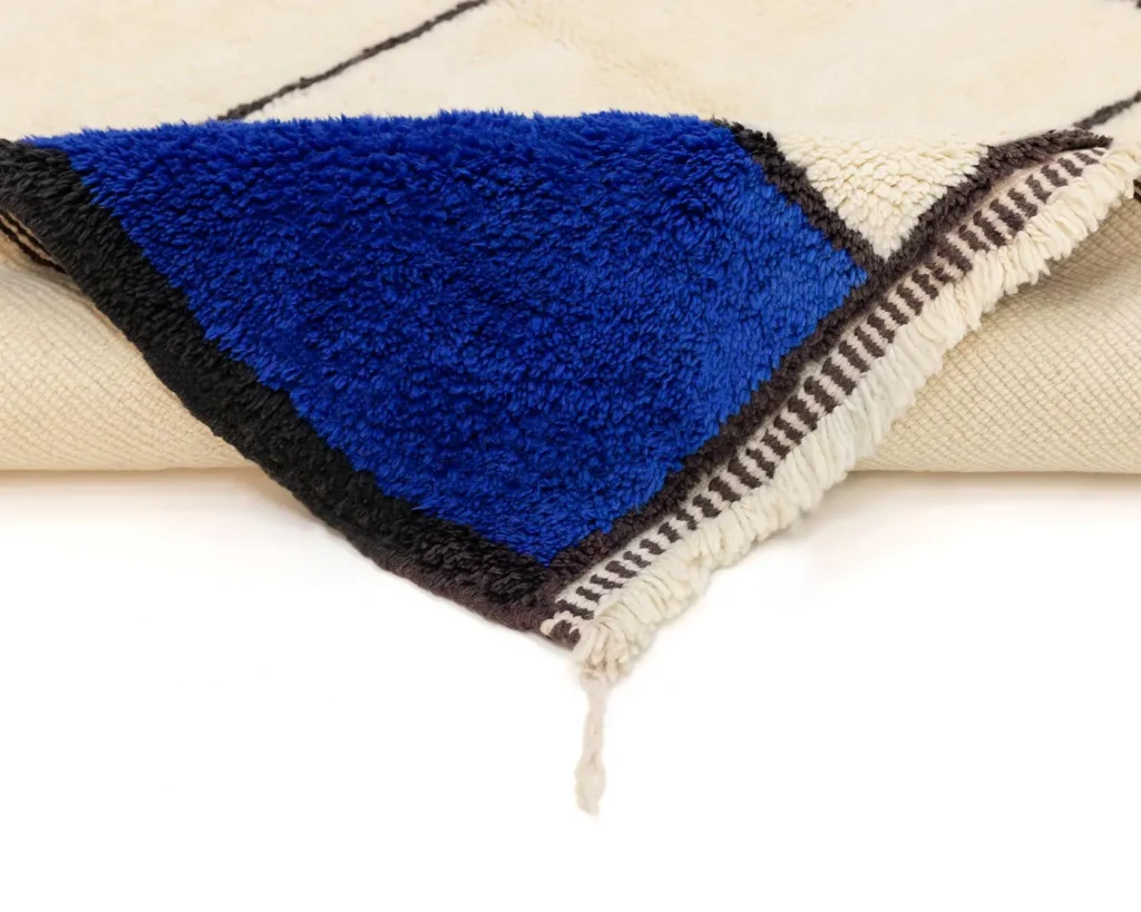 A corner of an authentic Moroccan rug with a blue, black, and white pattern is folded over, revealing its textured design and tassel edge.