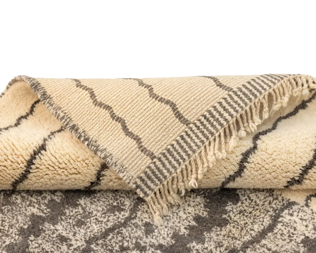A rolled-up beige and gray hand-knotted rug with fringe on one end, partially unfurled to showcase its texture and design.