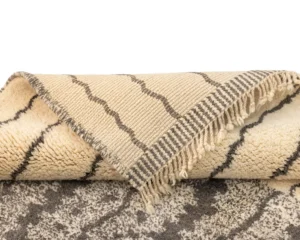 A rolled-up beige and gray hand-knotted rug with fringe on one end, partially unfurled to showcase its texture and design.