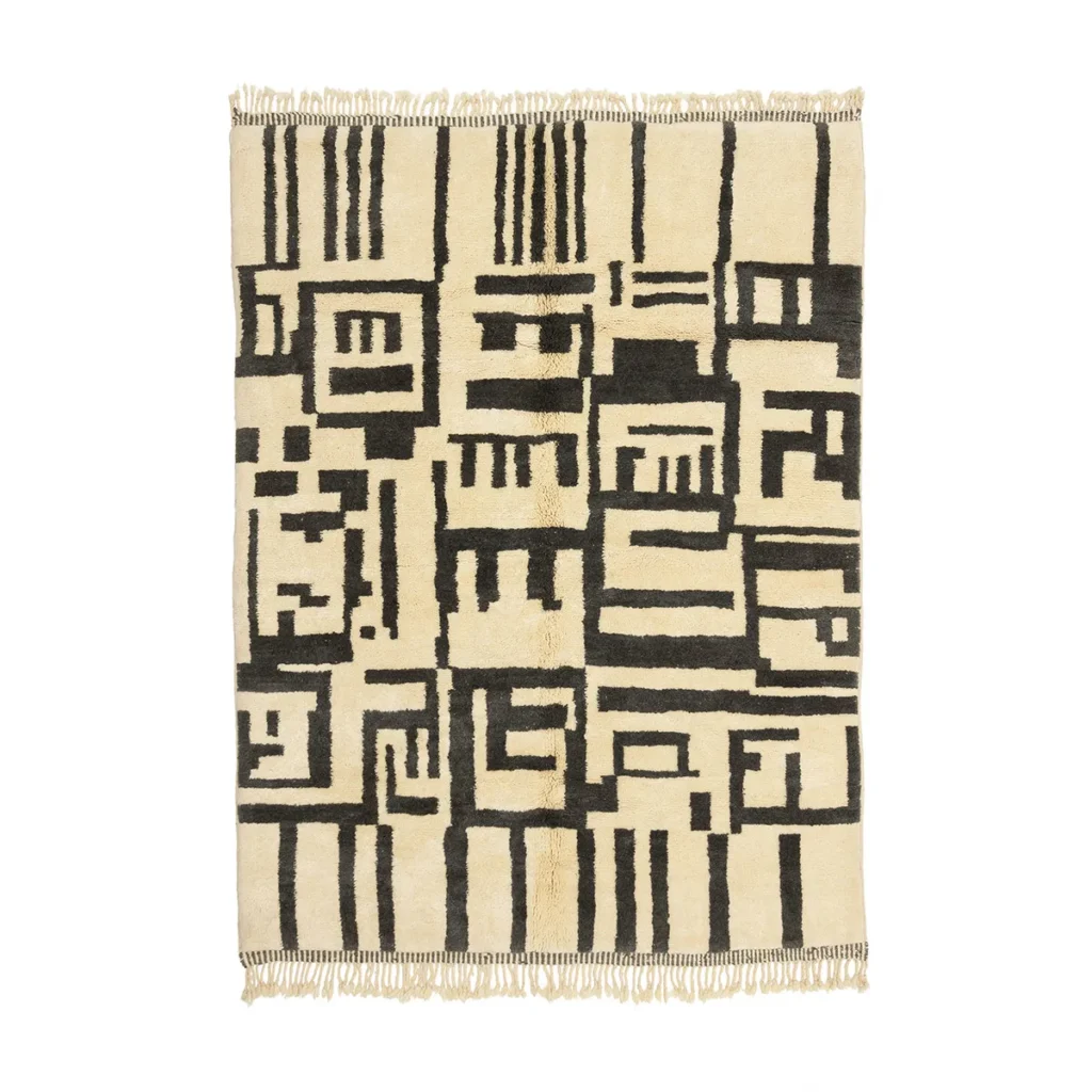 This handmade home decor piece is a cream and black rectangular rug showcasing an abstract geometric pattern with bold lines and shapes, complemented by fringed edges.