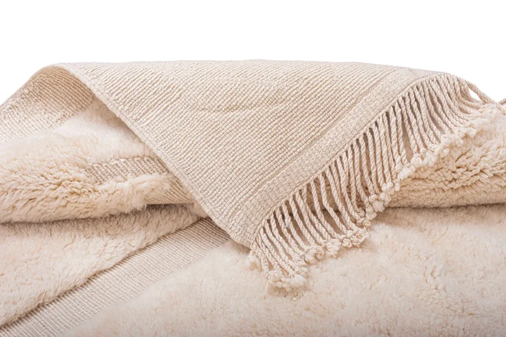 A hand-knotted beige fringed piece is elegantly laid over a fluffy cream-colored surface, echoing the essence of authentic Moroccan rugs with its artisanal charm.