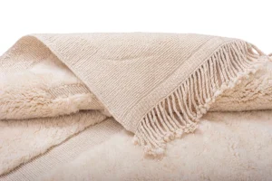 A hand-knotted beige fringed piece is elegantly laid over a fluffy cream-colored surface, echoing the essence of authentic Moroccan rugs with its artisanal charm.