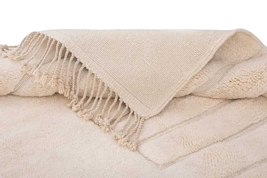 A beautifully crafted piece of handmade home decor, this beige textured wool creation features a fringed edge and is folded neatly on a harmoniously colored surface.