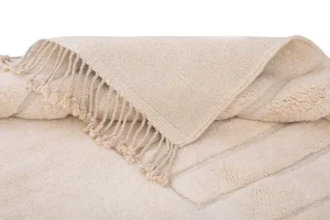 A beautifully crafted piece of handmade home decor, this beige textured wool creation features a fringed edge and is folded neatly on a harmoniously colored surface.