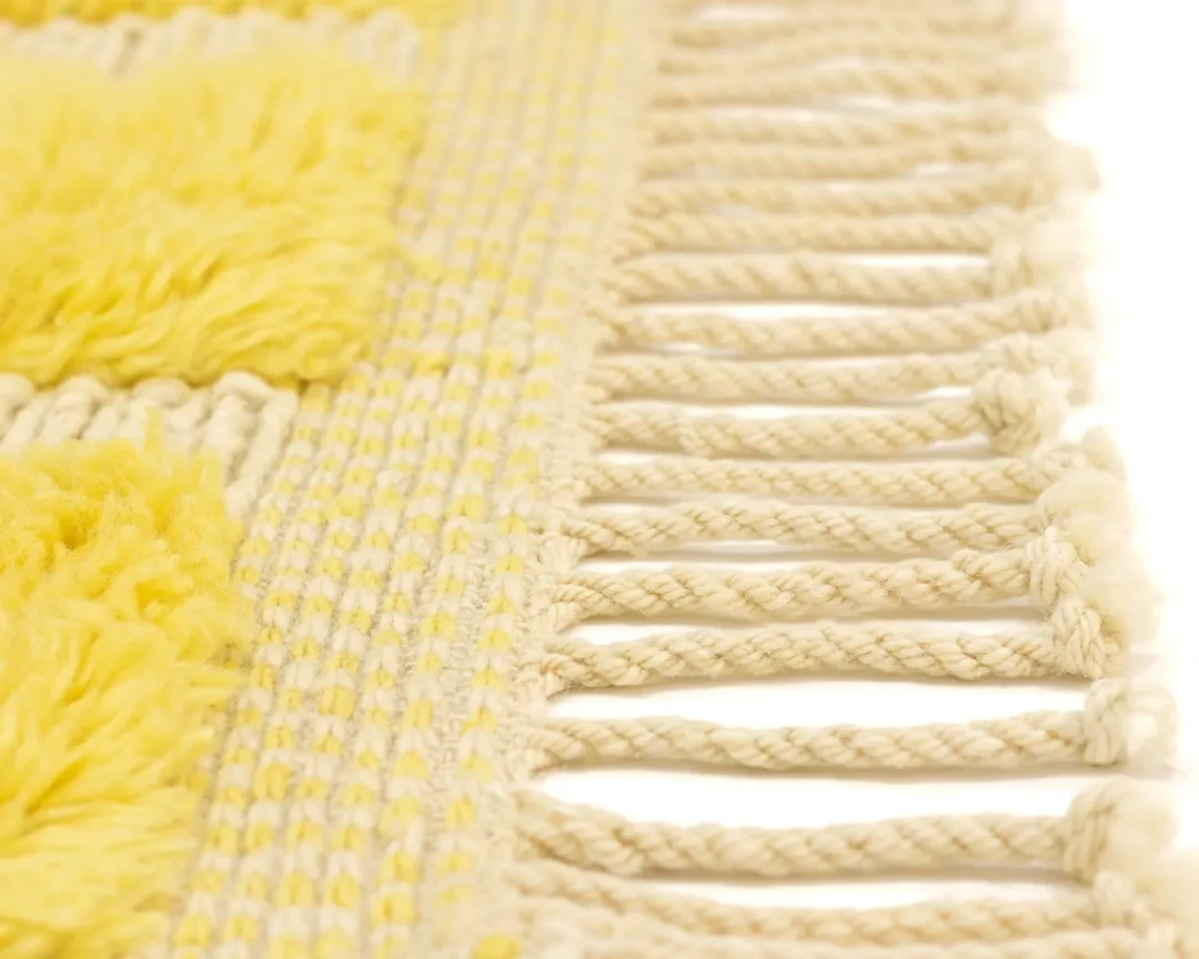 This close-up showcases a luxurious woven textile with a yellow and beige pattern, accentuated by braided beige fringe strands on the right edge. Perfectly embodying the elegance of handmade home decor, it stands as a testament to exquisite craftsmanship.