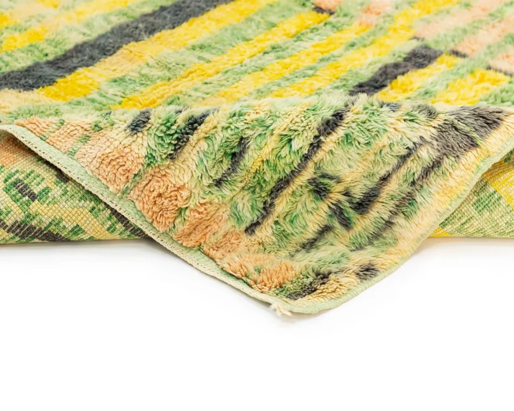 A close-up of a multicolored, textured artisanal rug reveals vibrant green, yellow, orange, and black stripes. The folded edges showcase a unique pattern on the underside, highlighting its hand-knotted craftsmanship.