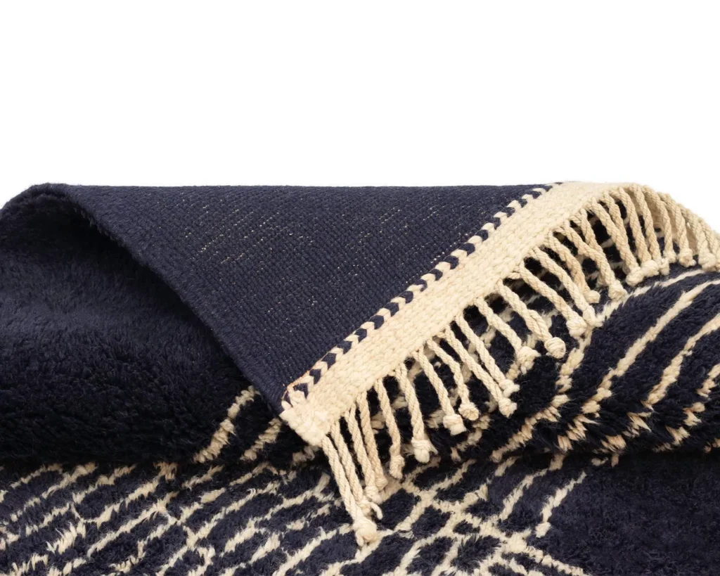 A beautifully crafted piece is displayed with fringed edges and a textured, patterned design in navy and cream. This luxurious item evokes the artistry found in hand-knotted rugs, adding sophistication to any room.