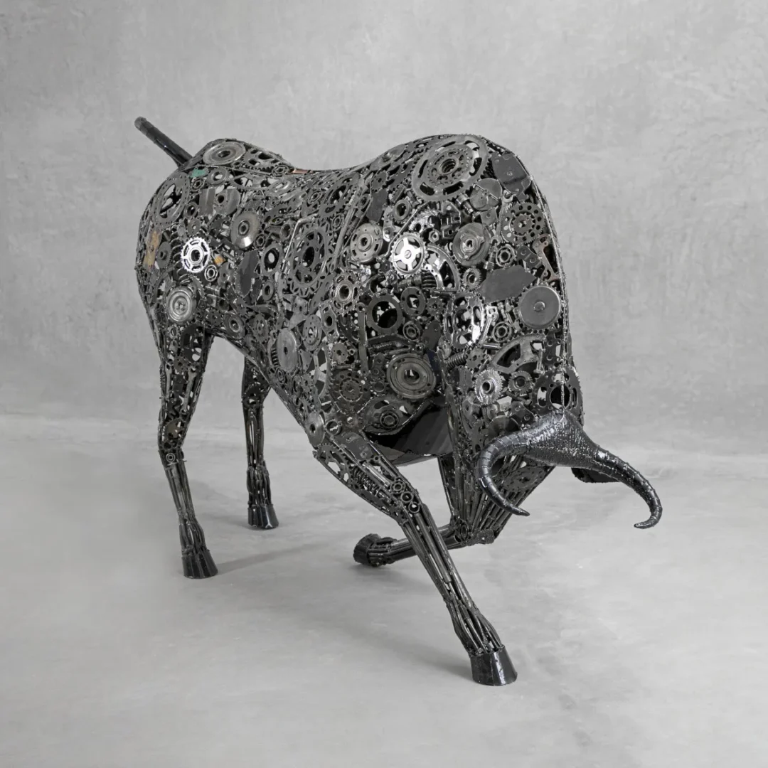 A Recycled Bull sculpture, crafted from metal gears and parts, stands in a charging pose against a gray background.