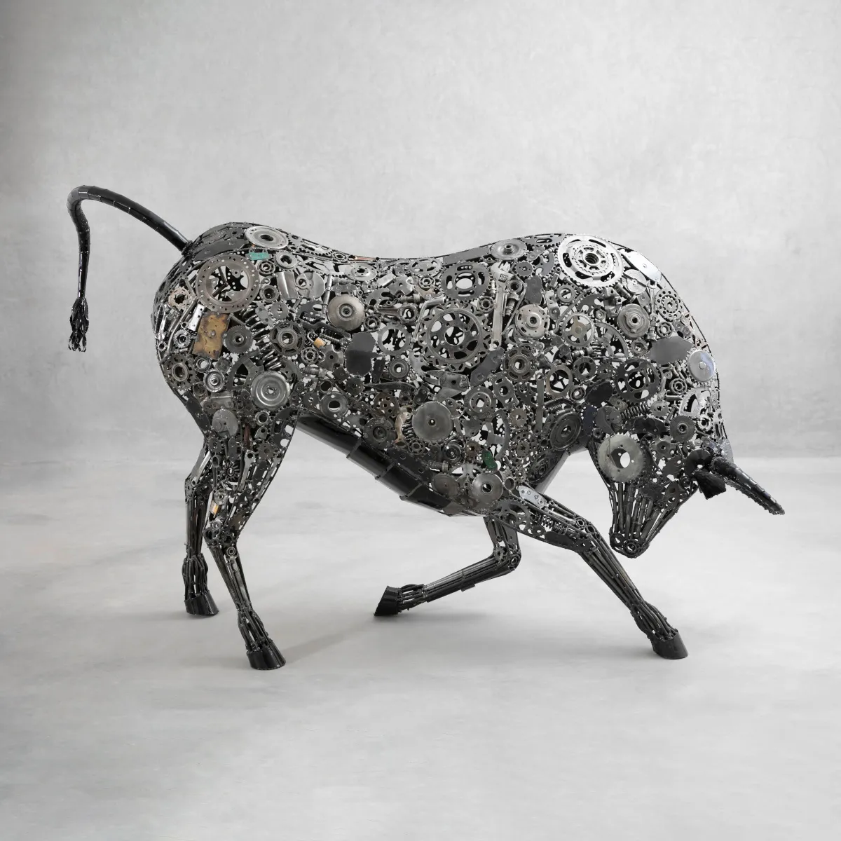 A bull sculpture made of various metal gears and parts stands on a light gray surface against a plain background.