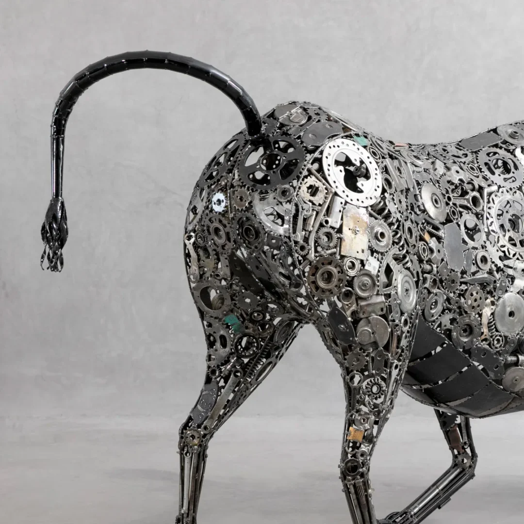 Recycled bull sculpture crafted from assorted gears and mechanical parts, showcasing the animals rear against a plain gray background; a fine example of creative metalwork.