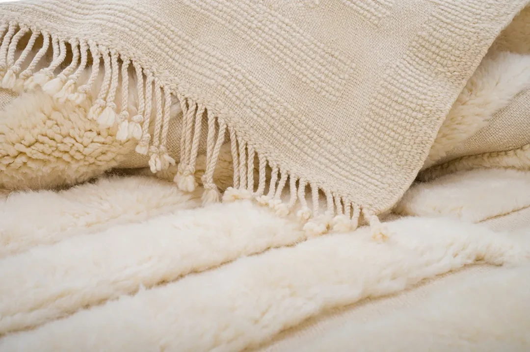 A close-up of a beige woven blanket with fringed edges draped over a soft, fluffy white surface.