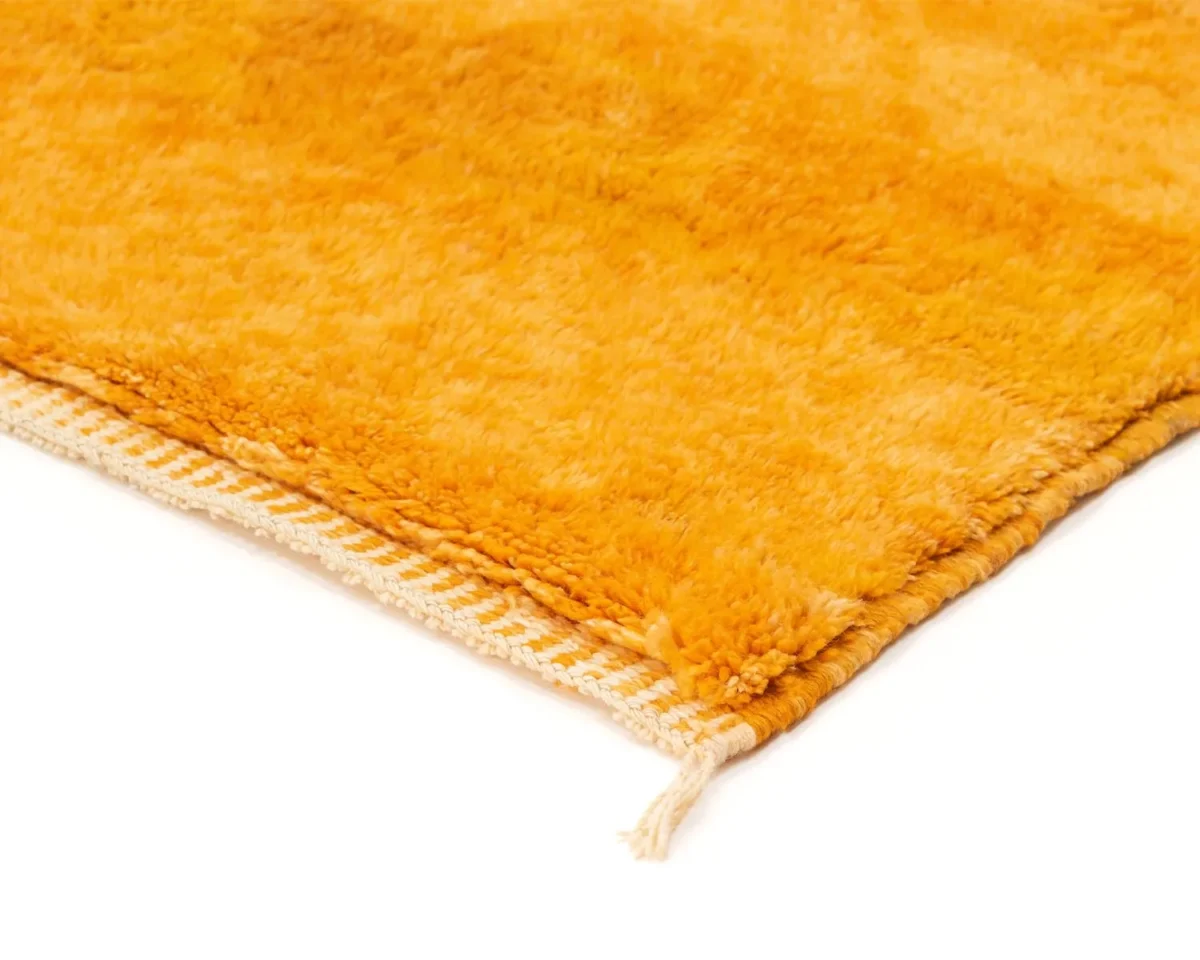 Close-up of a corner of an orange shag rug with thick pile and fringed edges on a white background.