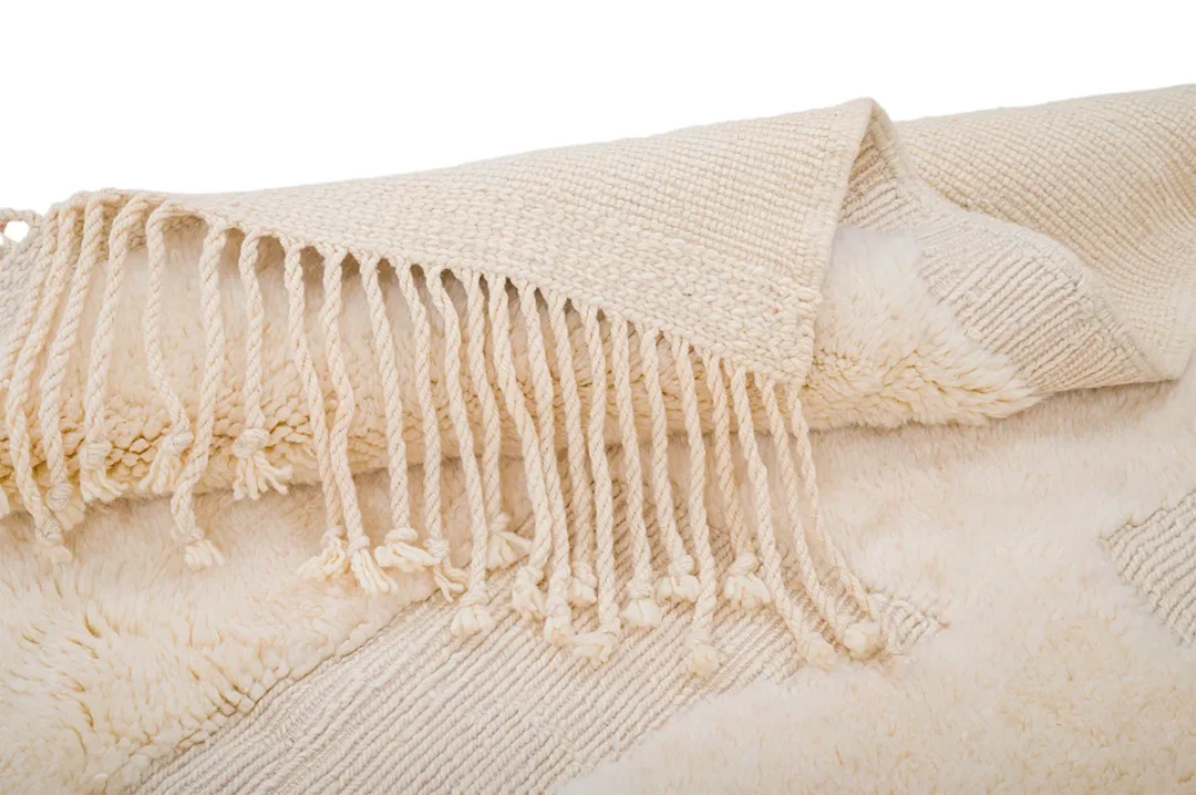 Close-up of a beige woven throw blanket with fringe tassels, draped over a textured surface.