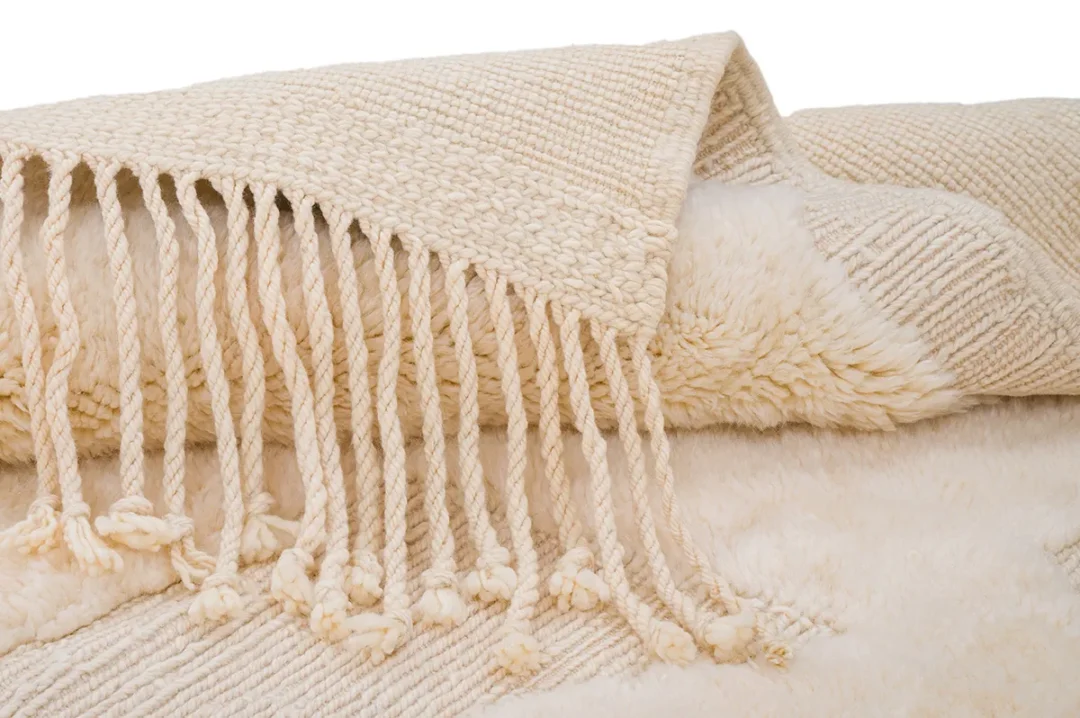 Beige woven throw blanket with fringe draped over a fluffy cream-colored surface.