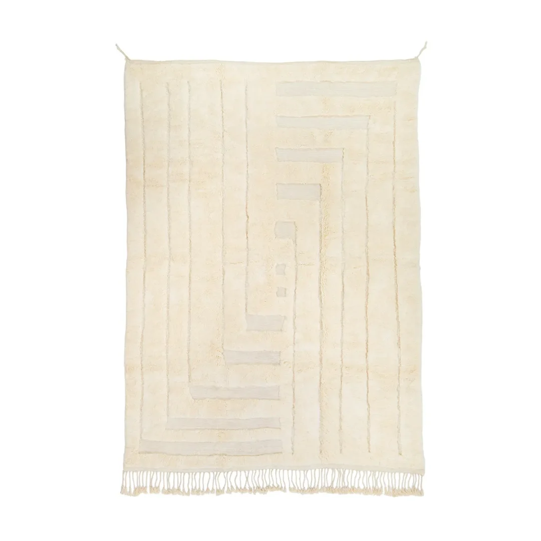 A beige geometric-patterned rug with vertical lines and shapes. It features fringe at the bottom edges, hanging against a plain white background.