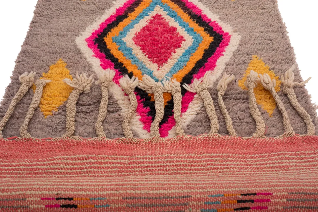 A multicolored woven rug with diamond and triangular patterns features fringe along the edge.