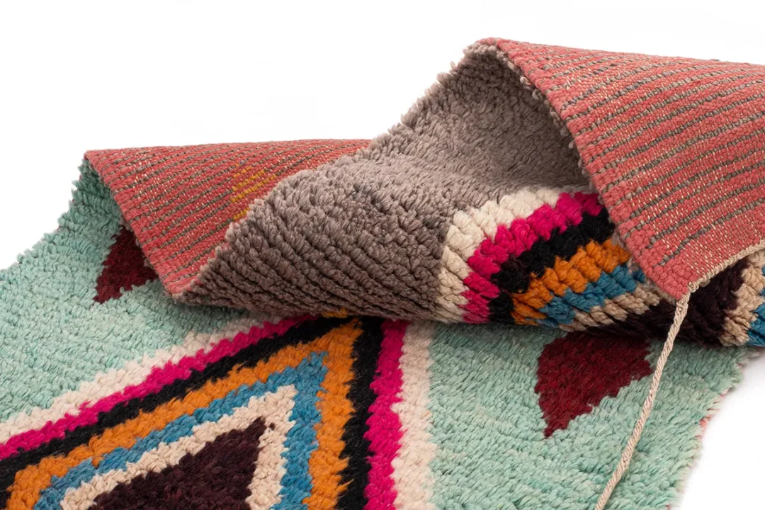 Colorful woven rug partially folded, showing a geometric pattern with red, blue, yellow, and black shapes on one side and a textured, gray-brown backing on the other.